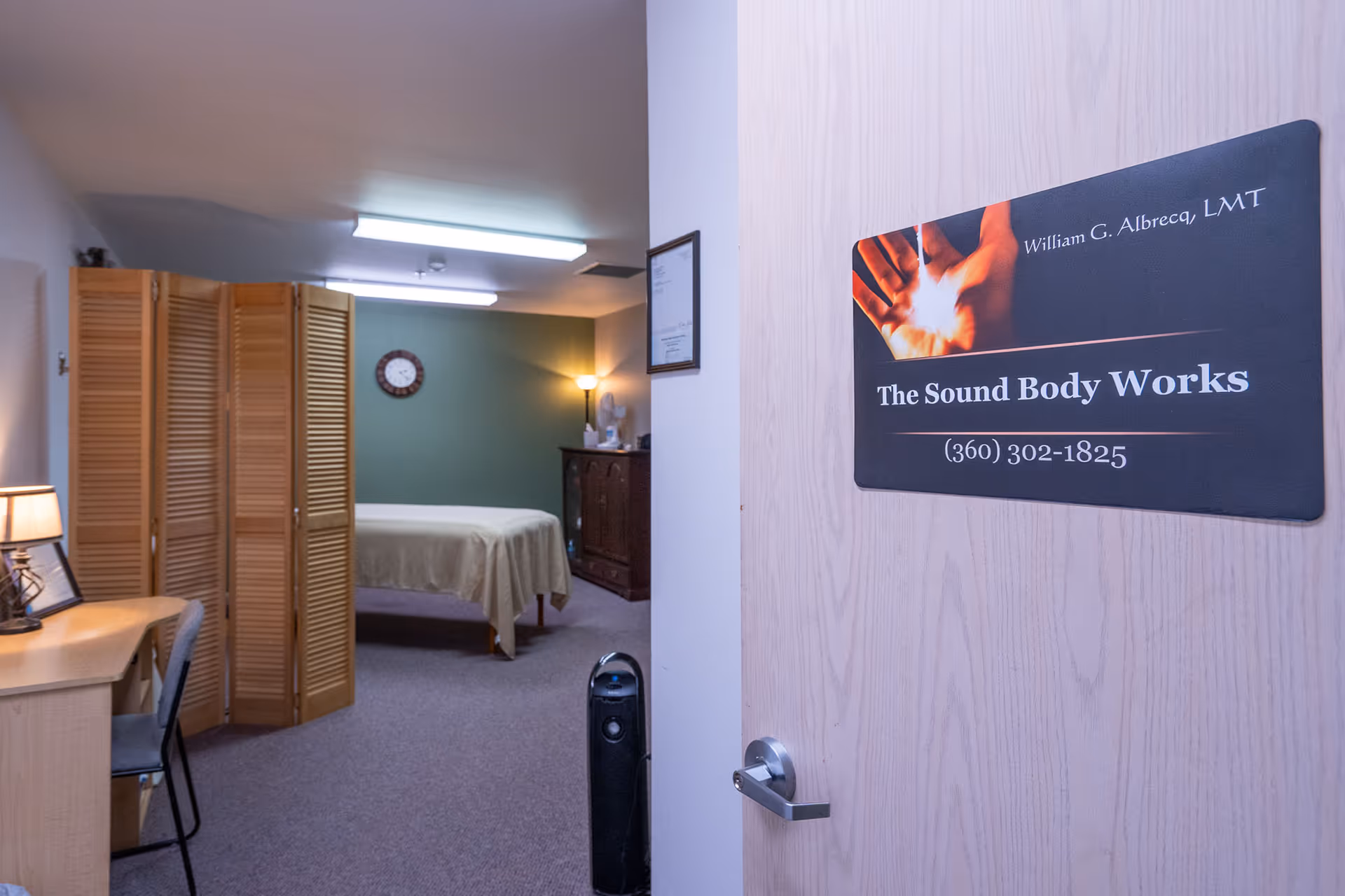 View into a softly lit room with a massage table covered in a beige sheet, a wooden folding screen, a desk with a chair and lamp, a clock on a green wall, and a wooden cabinet with a lamp and fan. The door has a sign that reads 'The Sound Body Works' with contact information.