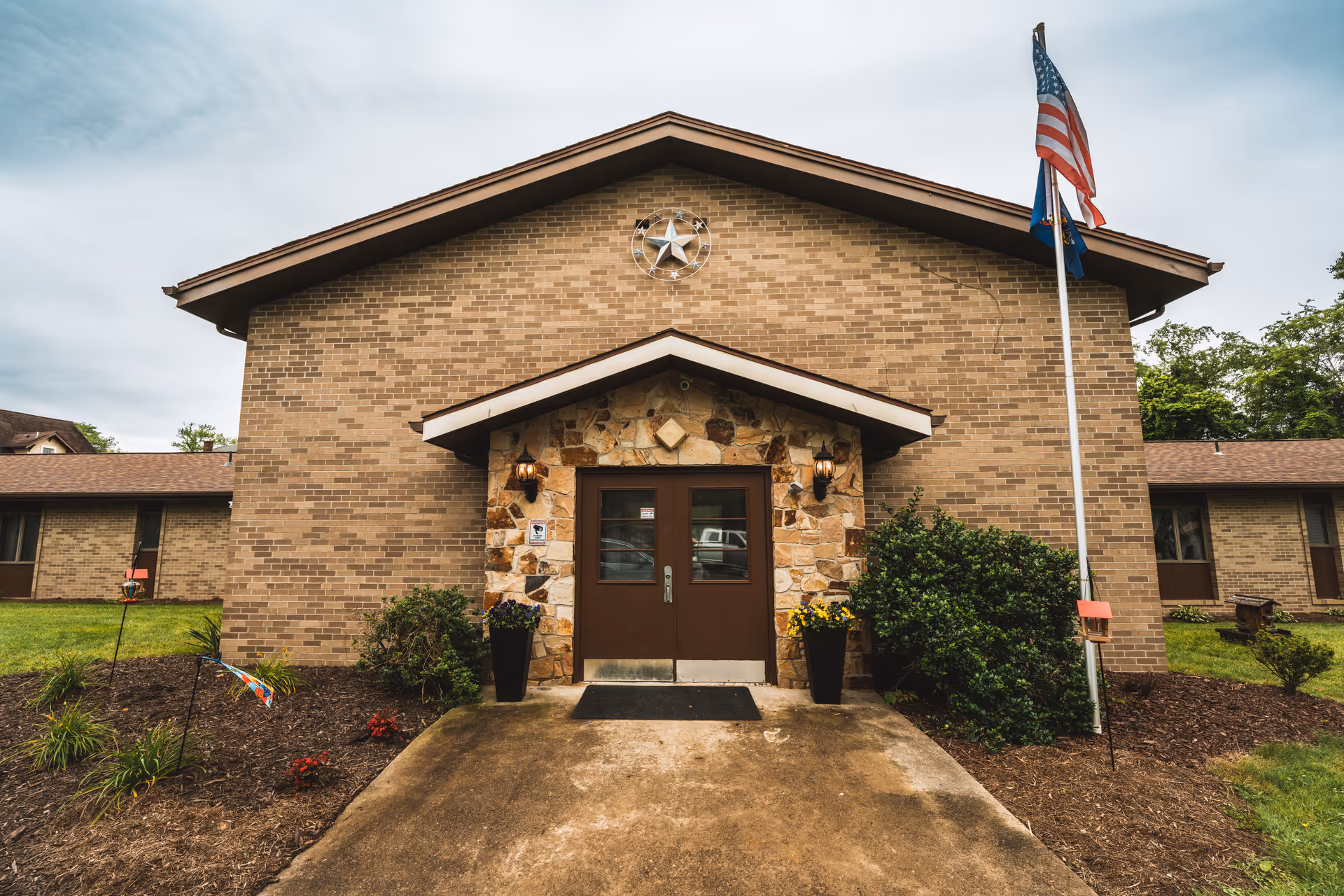 Front exterior view of a single-story brick building with a peaked roof and a stone entrance. Two brown doors are centered under a small overhang with two wall-mounted lanterns. There are two tall black planters with flowers on either side of the entrance. An American flag and another flag are flying on a flagpole to the right of the entrance. The building is surrounded by grass and some small shrubs and plants.