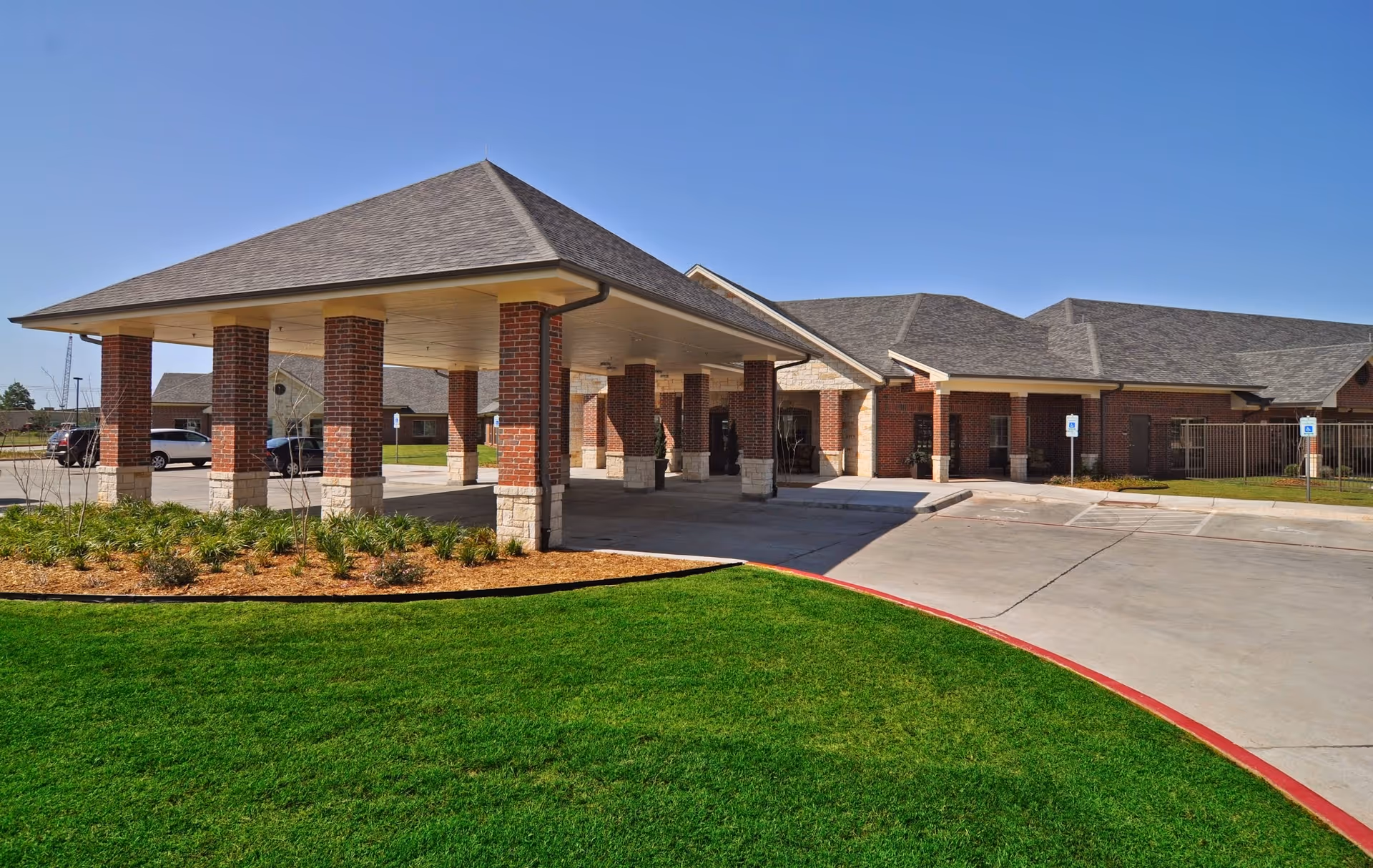 Exterior view of Medical Park West Rehabilitation and Skilled Care building with a covered entrance supported by brick columns, a driveway, and well-maintained green lawn under a clear blue sky.