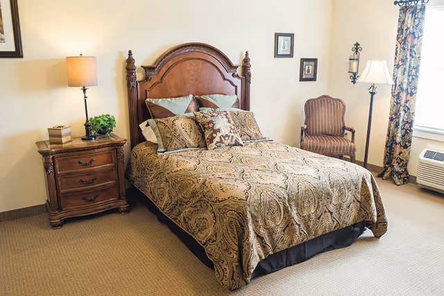 Bedroom featuring a wooden headboard bed with patterned bedding, a nightstand and lamp, and a striped armchair by the window.