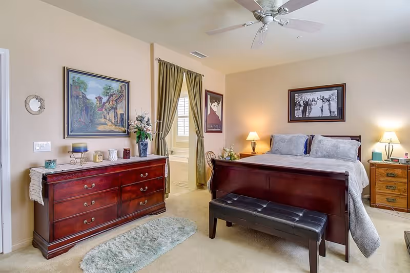A well-lit bedroom featuring a large wooden bed with gray bedding and pillows, two bedside tables with lamps, a wooden dresser with decorative items, a small bench at the foot of the bed, a green curtain partially covering a doorway leading to a bathroom, and framed artwork on the walls.