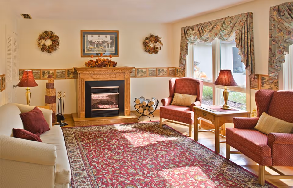 A cozy living room with a white sofa and two red armchairs, each with a beige pillow. There is a wooden fireplace with decorative wreaths and a framed picture above it. A patterned red area rug covers the floor, and two table lamps with red shades sit on wooden side tables. Large windows with floral curtains let in natural light, and a log holder with firewood is placed next to the fireplace.