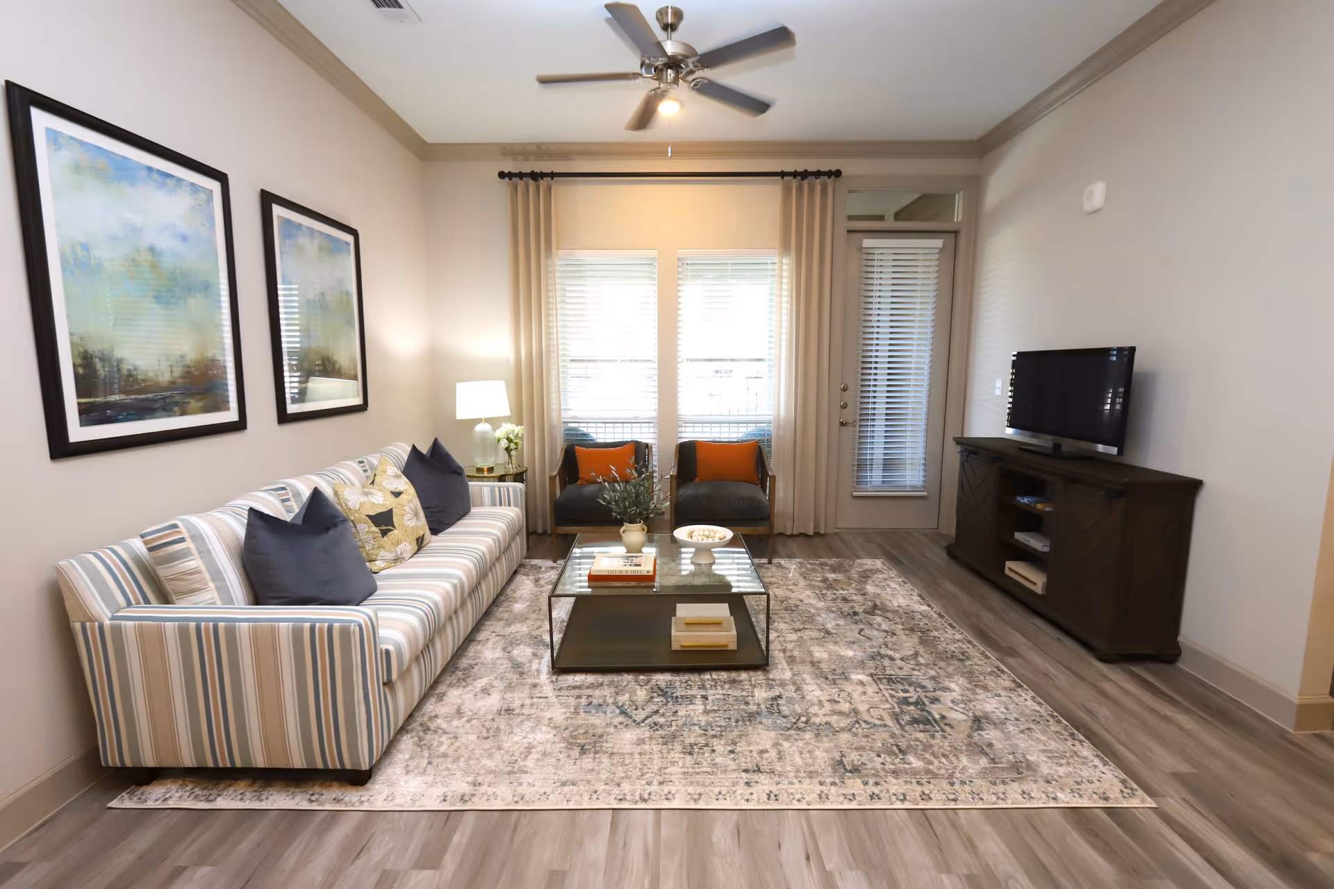 A bright living room with a striped sofa adorned with dark and floral pillows, two armchairs with orange cushions, a glass coffee table with decorative items, a patterned area rug, a ceiling fan with light, two framed abstract paintings on the wall, a floor lamp, and a TV on a wooden stand. There is a window with blinds and curtains, and a door with blinds next to it.