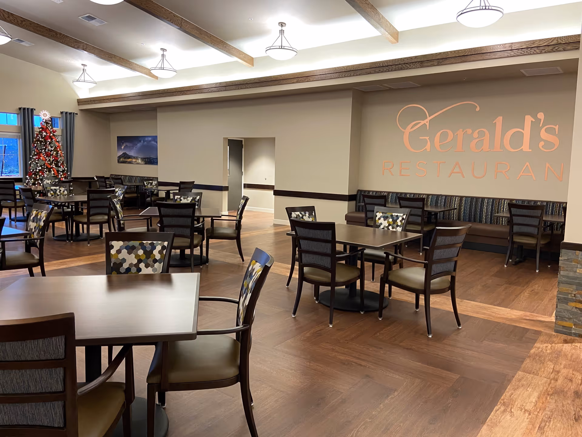 Interior view of Gerald's Restaurant at Fieldstone on Clear Creek, featuring multiple dining tables and chairs with patterned upholstery, wooden floors, exposed wooden ceiling beams, and a decorated Christmas tree near the windows.