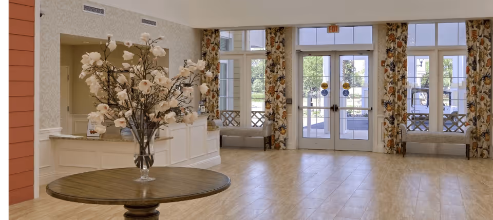 Spacious and well-lit lobby area with a round wooden table in the center holding a large vase of white flowers. The background features large glass doors and windows with floral curtains, two cushioned benches, and a reception desk to the left.