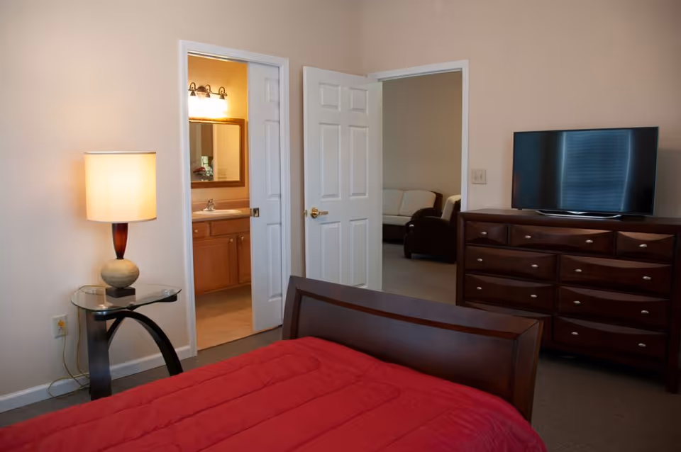 A bedroom with a wooden bed frame and a red comforter. To the left is a glass-top side table with a lamp. Two open doors reveal a bathroom with a sink and mirror, and a living area with a sofa. A wooden dresser with a flat-screen TV on top is on the right side of the room.