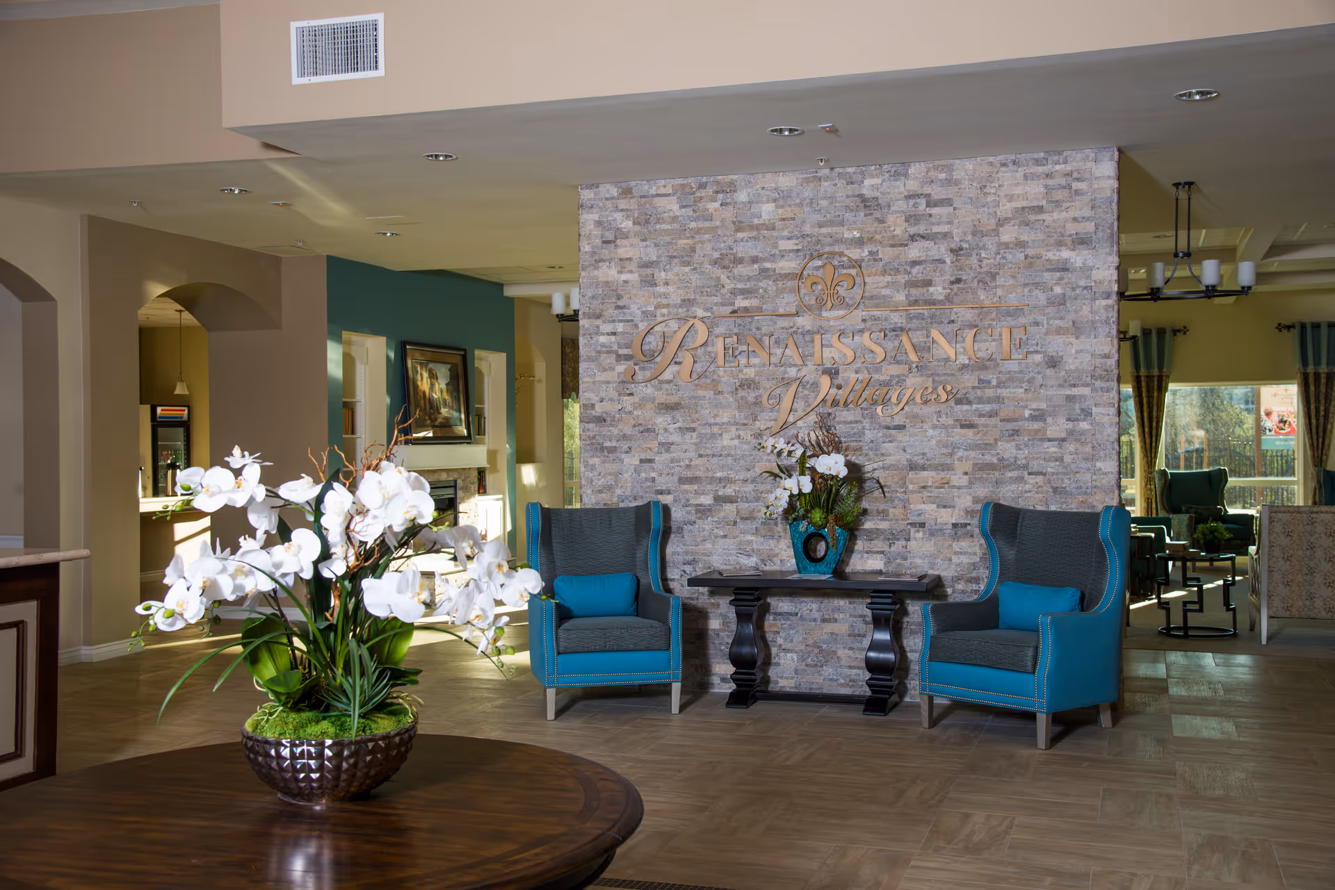 Lobby area of Renaissance Villages featuring a stone accent wall with the facility's name, two teal armchairs with blue cushions, a dark wooden table with a floral arrangement, and a spacious interior with natural light.