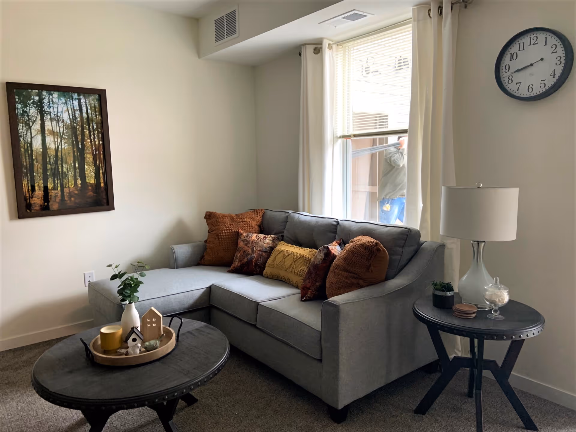 A cozy living room with a gray sectional sofa adorned with multiple decorative pillows in shades of brown and orange. A round black coffee table in front of the sofa holds a tray with small decorative items including a white vase with greenery, a small house figurine, and a candle. To the right of the sofa is a round side table with a white lamp, a small plant, and a glass container. A large window with white curtains lets in natural light, and a wall clock is mounted above the side table. A framed picture of a forest scene hangs on the wall to the left.