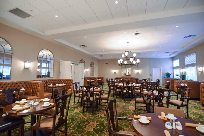 A spacious dining room with multiple round wooden tables and chairs arranged neatly. The tables are set with white cups, saucers, and napkins. The room features tufted leather banquette seating along the walls, large mirrors, wall sconces, and a central chandelier. The carpet has a green and gold pattern, and windows with blinds allow natural light to enter.