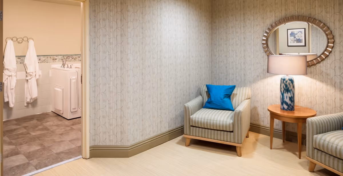 A cozy sitting area with two striped armchairs, each with a blue cushion, and a wooden side table with a blue and white patterned lamp. An oval mirror hangs on the wallpapered wall above the table. To the left, an open doorway reveals a bathroom with two white robes hanging on a towel rack and a walk-in bathtub.