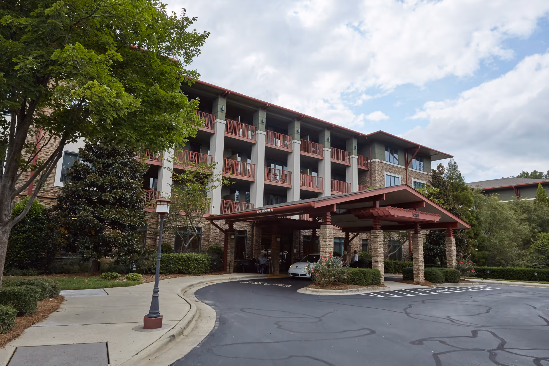 Exterior view of a multi-story senior living facility named Magnolia Glen with a covered entrance, stone pillars, balconies, and surrounding greenery under a partly cloudy sky.