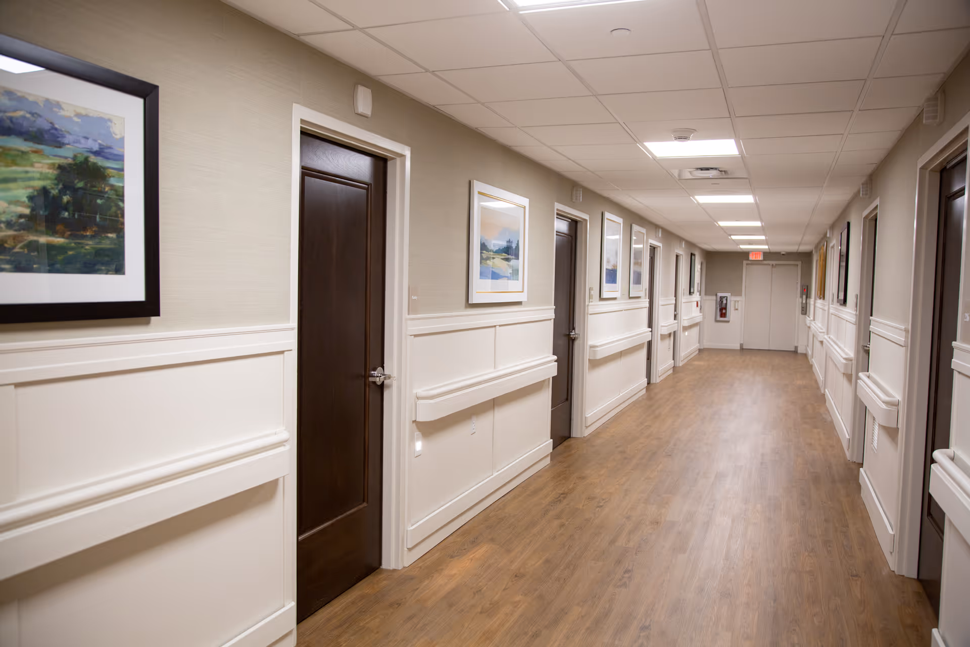A long, well-lit hallway in a senior living facility with wooden doors on both sides, beige walls with white wainscoting, handrails, and framed landscape paintings hanging on the walls. The floor is wood-patterned, and there is a fire extinguisher mounted on the far wall near an exit door.