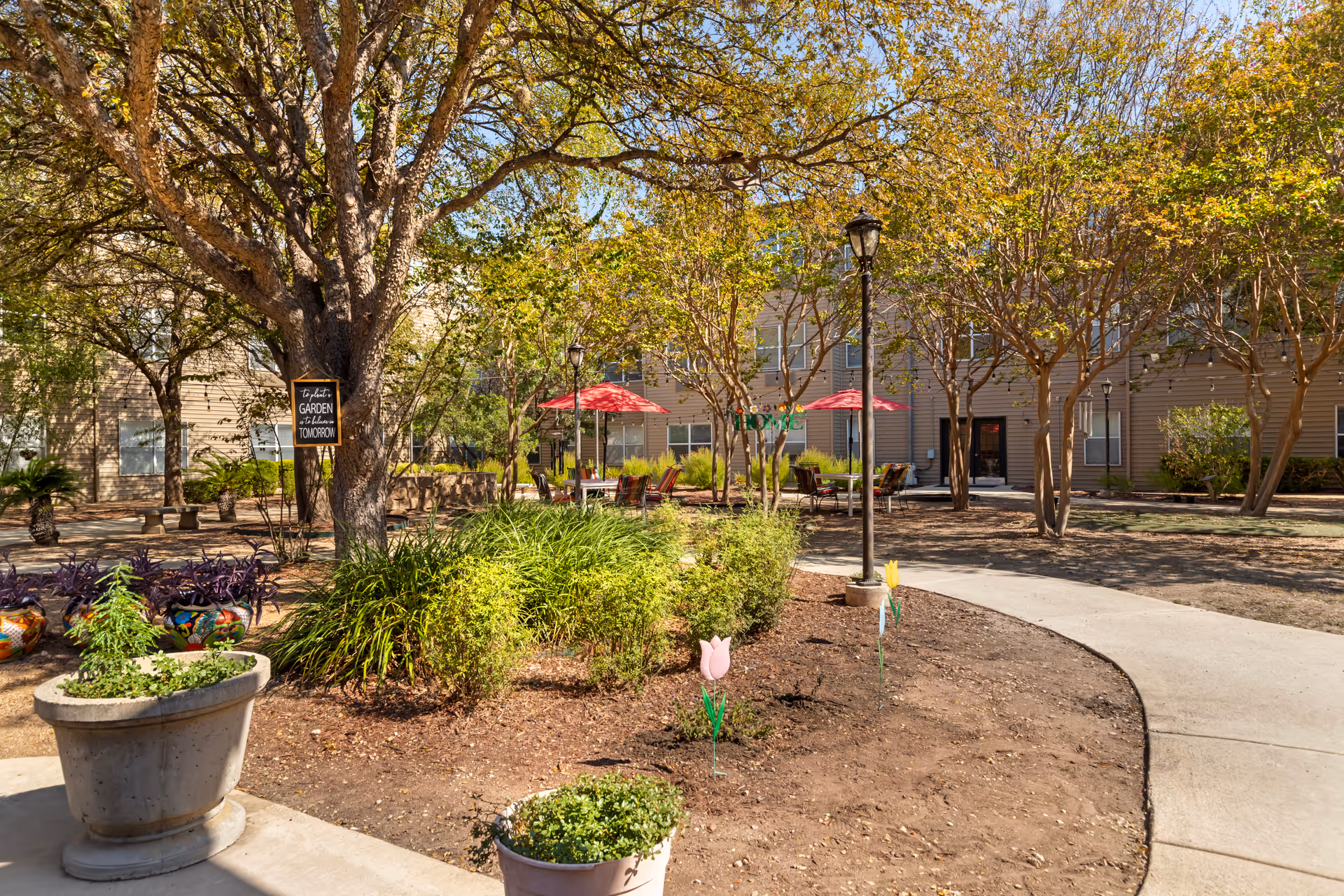 Sunny courtyard of a senior living facility with trees, potted plants, a curved walkway, and seating areas with red umbrellas in front of the building.