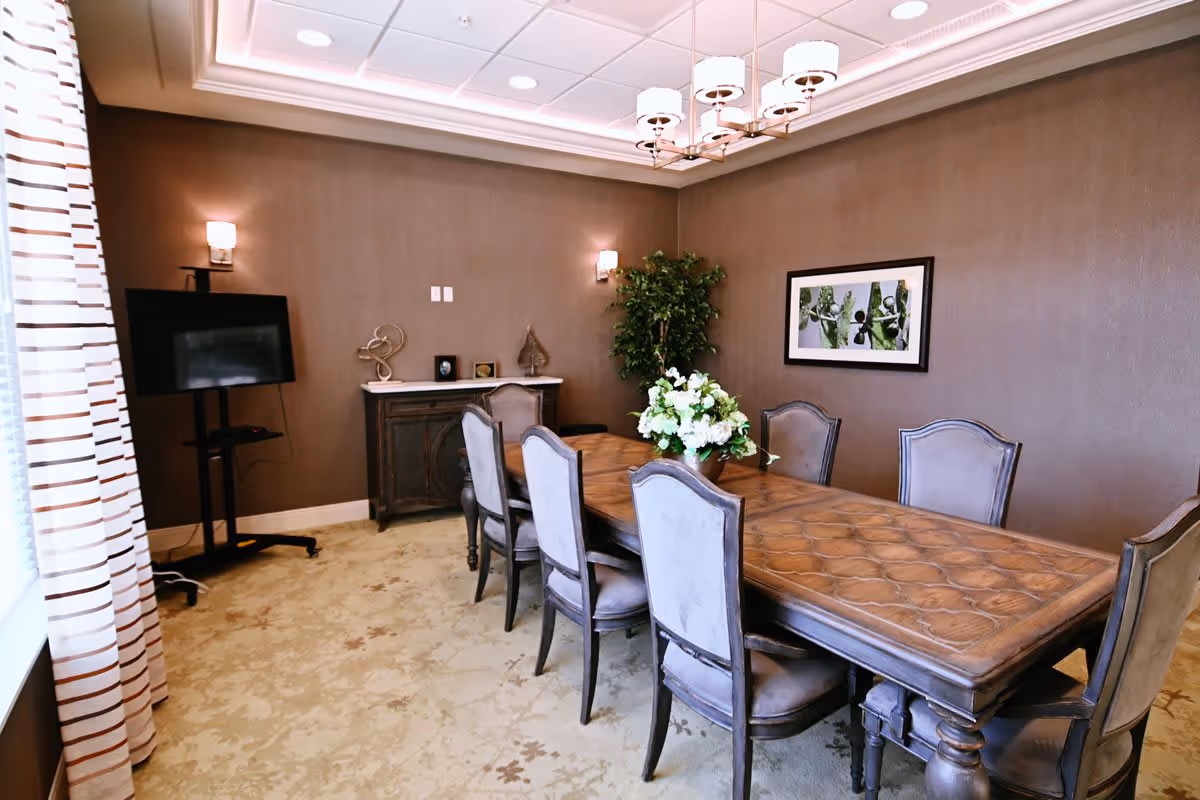 A formal dining room with a long wooden table surrounded by eight upholstered chairs. The room features brown walls, a patterned carpet, a sideboard with decorative items, a flat-screen TV on a stand, a large potted plant, and a framed picture on the wall. A modern chandelier hangs from the ceiling, and a window with striped curtains allows natural light into the room.