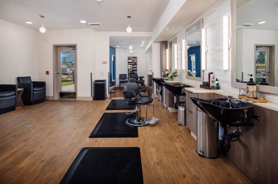 Bright modern salon with styling chairs, shampoo sinks, mirrors, and a seating area in a senior living facility.