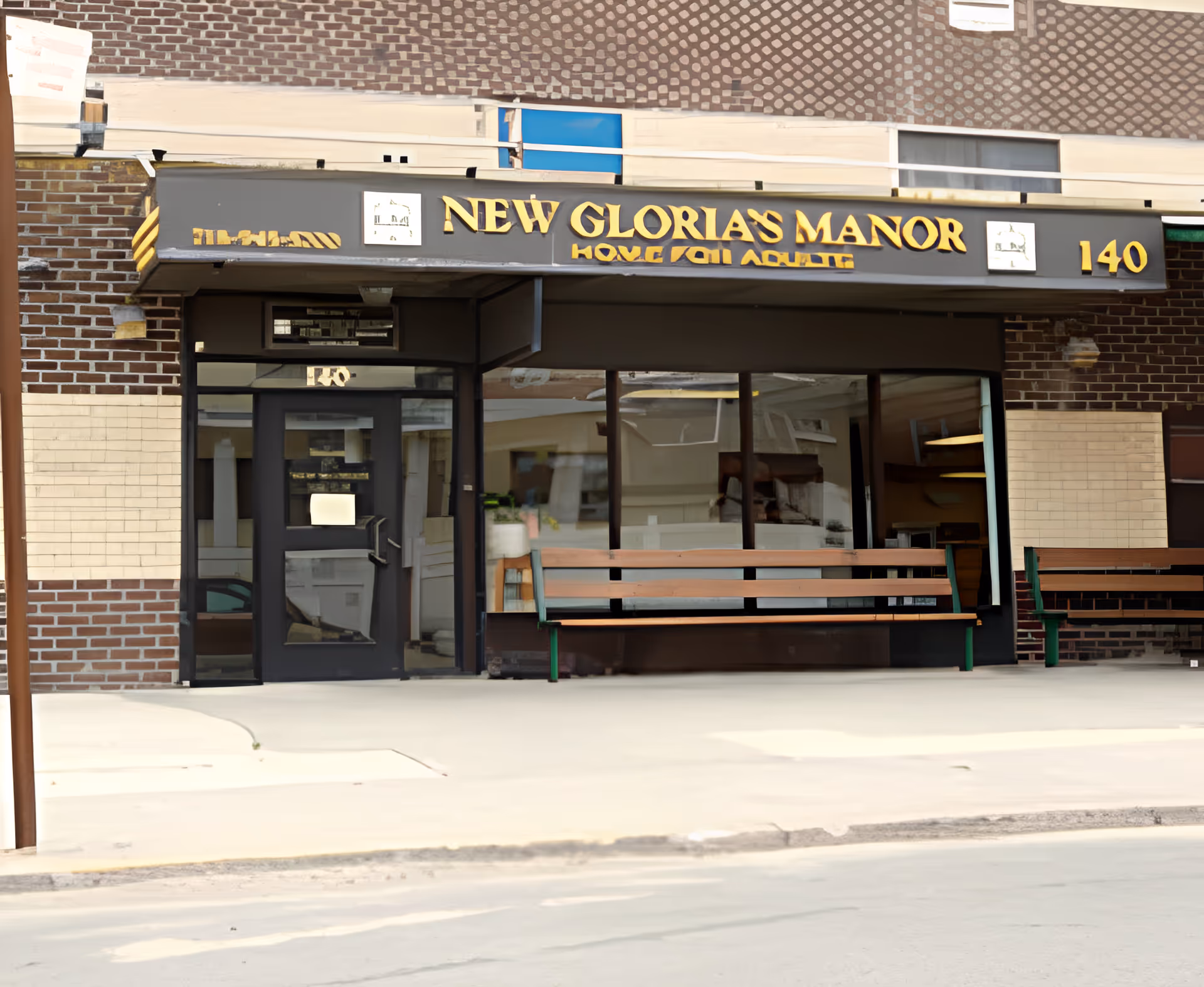 Front exterior view of New Glorias Manor Home For Adults building with a black entrance door, large windows, and two wooden benches outside on the sidewalk. The building has a brick facade and a sign above the entrance displaying the facility name and address number 140.