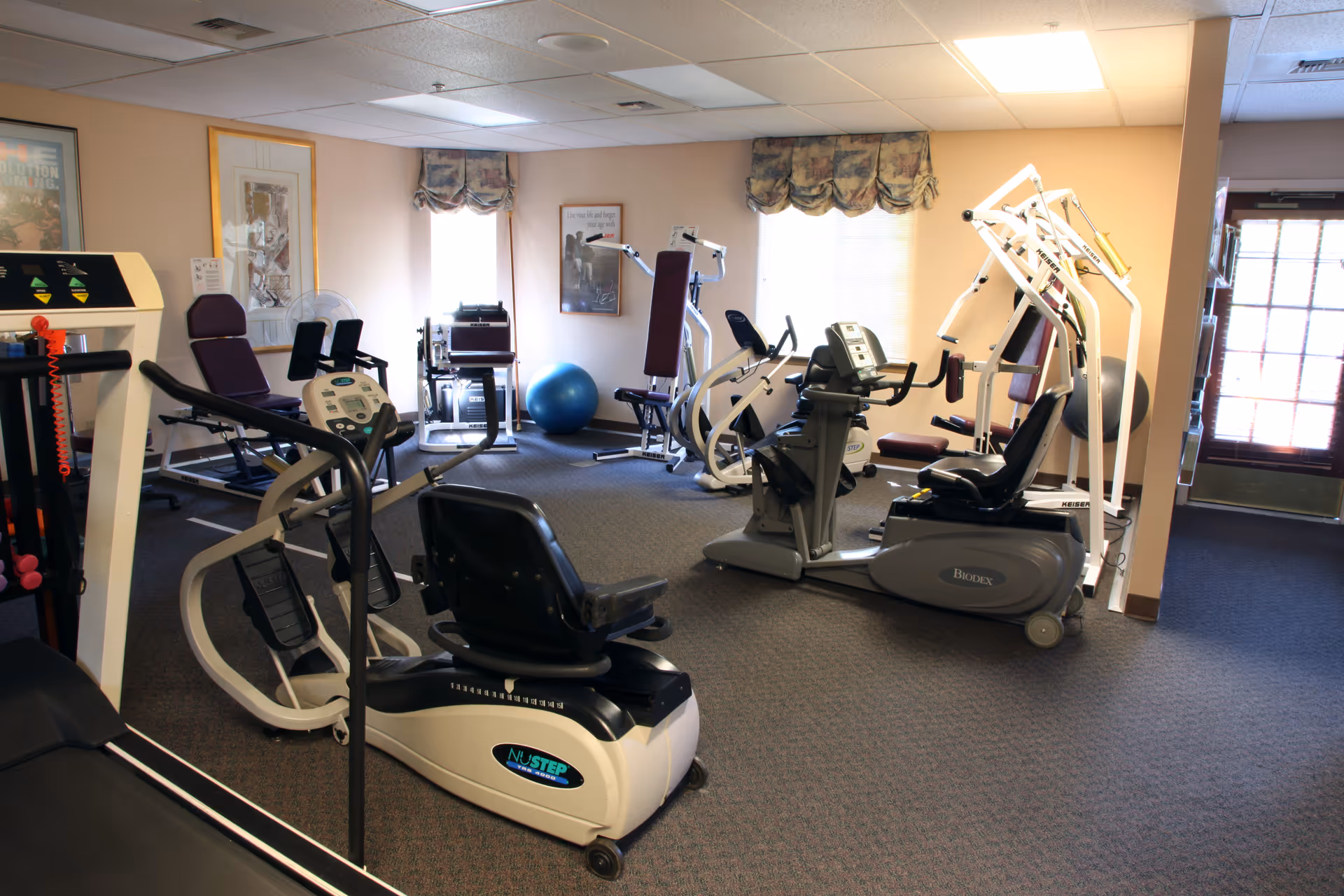 Indoor fitness room with various exercise equipment including stationary bikes, weight machines, and a blue exercise ball. The room has carpeted flooring, beige walls, two windows with floral valances, and framed pictures on the walls. There is a door with glass panels on the right side.