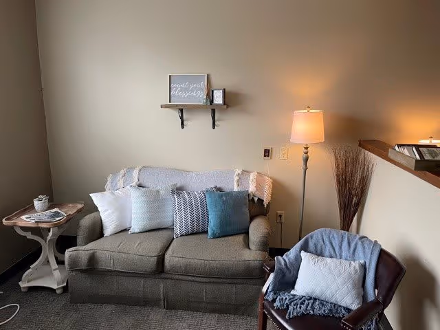 A cozy living room area with a gray loveseat adorned with five decorative pillows and a throw blanket. To the right, there is a dark brown armchair with a white pillow and a blue throw blanket. A floor lamp with a beige shade stands behind the armchair, casting warm light. On the left side, there is a small wooden side table with a potted plant and a magazine. Above the loveseat, a small wooden shelf holds a framed sign and a small picture frame. The walls are painted a light beige color.