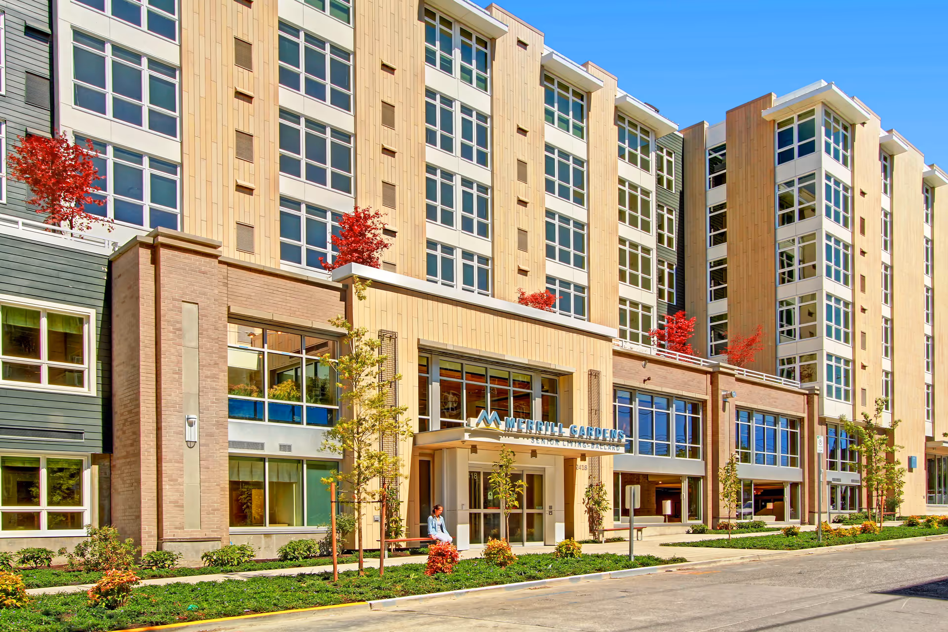 Exterior view of Merrill Gardens at Ballard, a multi-story senior living facility with large windows, beige and brown facade, small trees and shrubs in front, and a person sitting near the entrance.