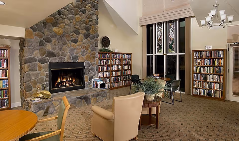 Cozy living room area with a large stone fireplace, bookshelves filled with books, a round wooden table with chairs, an armchair, and a small table with a potted plant. Large windows with decorative stained glass panels are visible in the background.