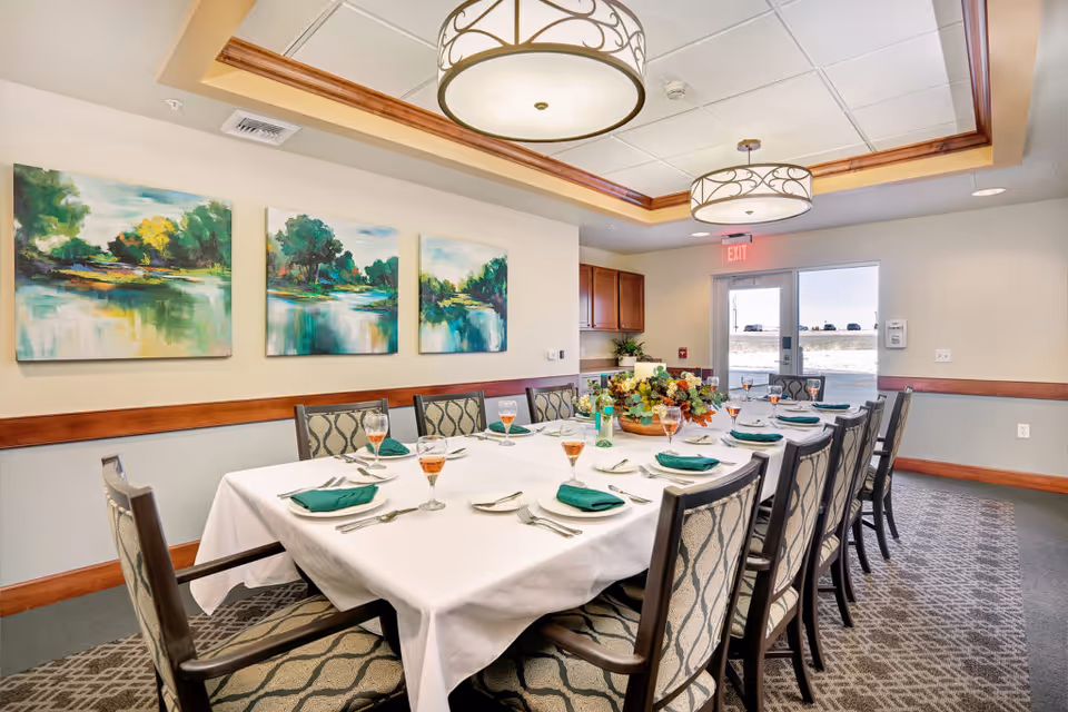 A formal dining room with a long rectangular table covered with a white tablecloth, set with green napkins, silverware, and glasses filled with a light beverage. The room features patterned carpet, cushioned chairs with wooden frames, three landscape paintings on the wall, two large decorative ceiling lights, and a glass door leading outside.