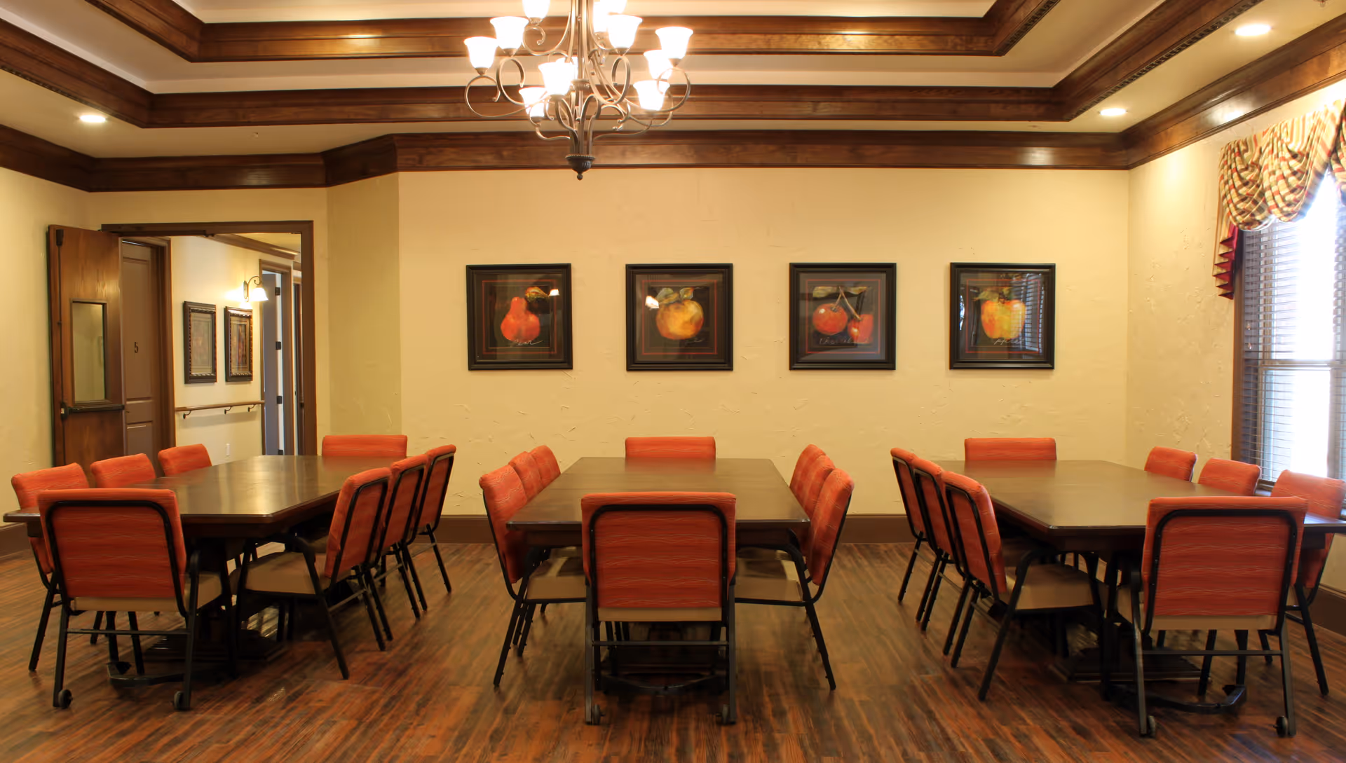 A dining room with three rectangular wooden tables, each surrounded by red cushioned chairs. The room has wooden flooring, cream-colored walls, and a coffered ceiling with a chandelier. Four framed pictures of fruit hang on the wall, and a window with patterned curtains is on the right side.