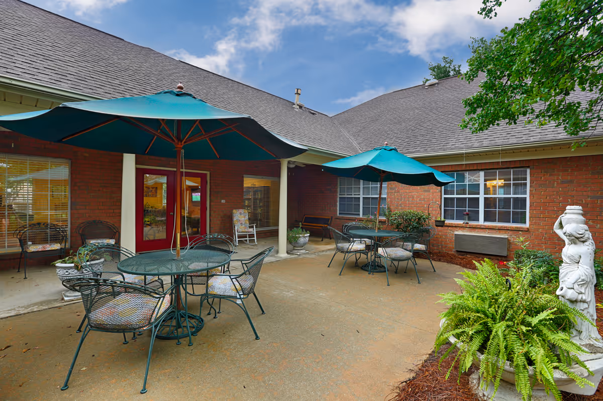 Outdoor patio area with two round metal tables, each with four chairs and a large green umbrella. The patio is adjacent to a brick building with windows and a red door. There are plants, including a large fern and a white statue, around the patio. The sky is partly cloudy.