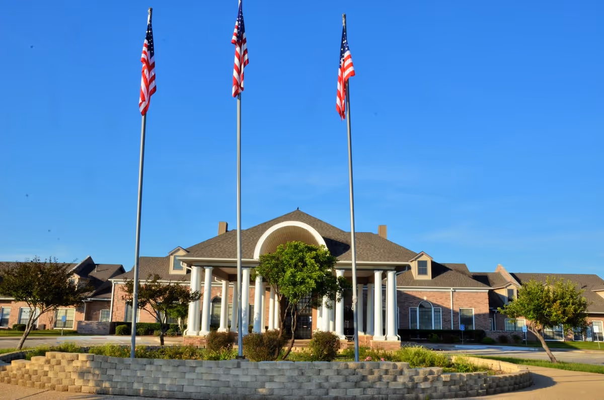 Front exterior view of Rosewood Villas building with three tall flagpoles displaying American flags, a circular driveway, and landscaping with trees and bushes under a clear blue sky.