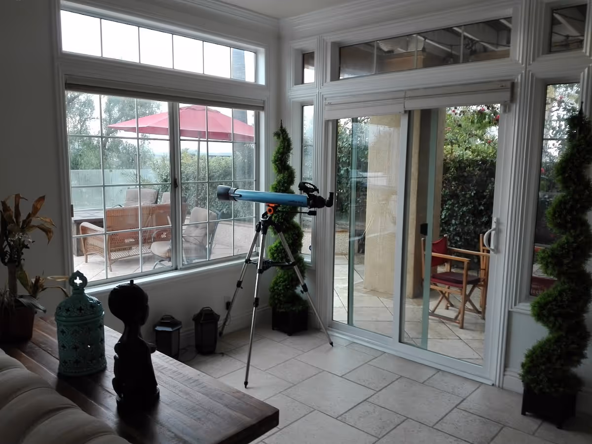 Interior view of a room with large windows and sliding glass doors leading to an outdoor patio. Inside, there is a telescope on a tripod positioned near the glass doors. The room has tiled flooring and decorative plants, with outdoor furniture visible through the windows and doors.