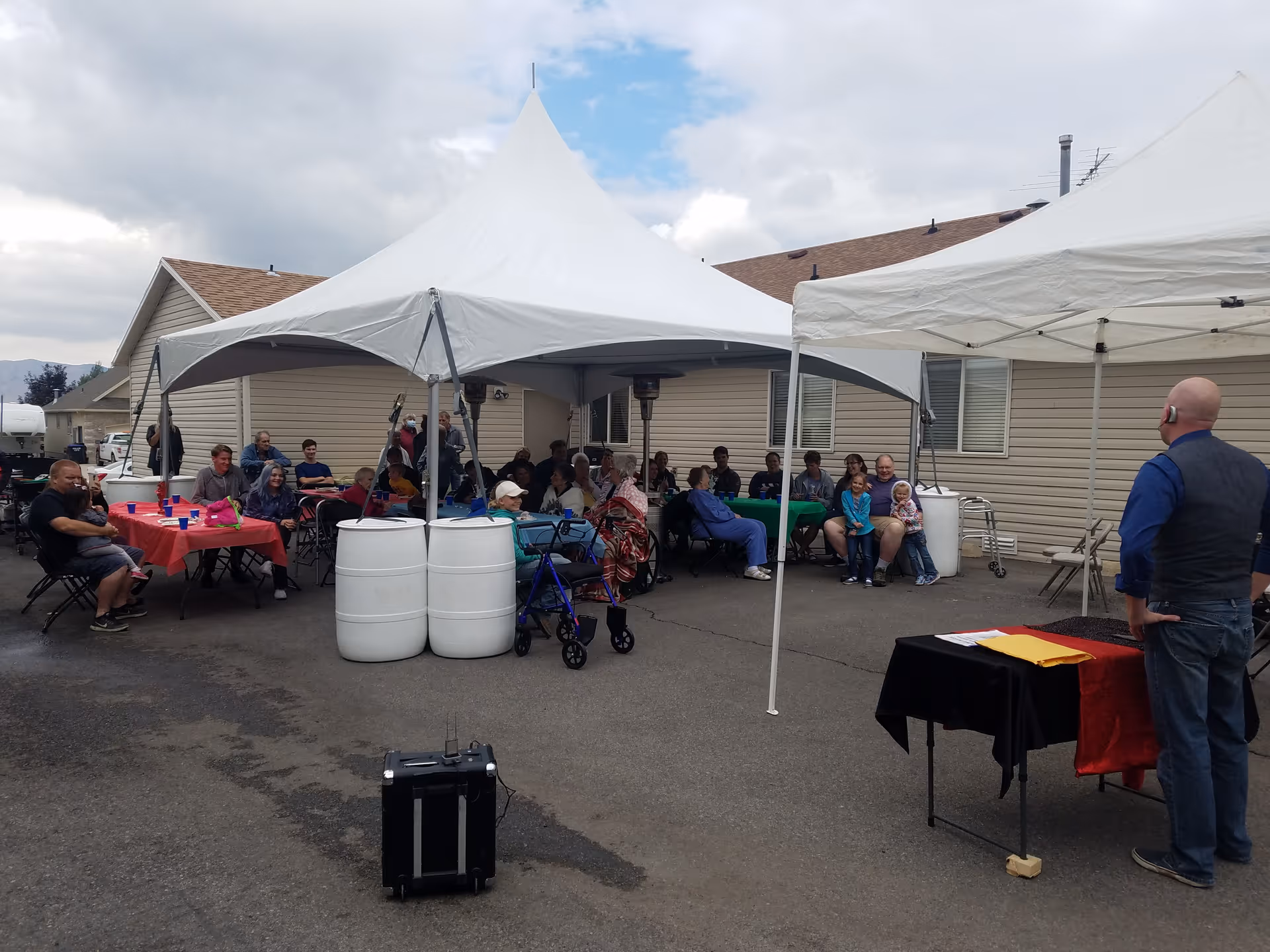 A group of people seated under two white canopy tents outside a beige building, with tables covered in red and green tablecloths. A man stands near a small table with a black and red cloth, facing the seated crowd. The setting appears to be a social gathering or event on a paved area.