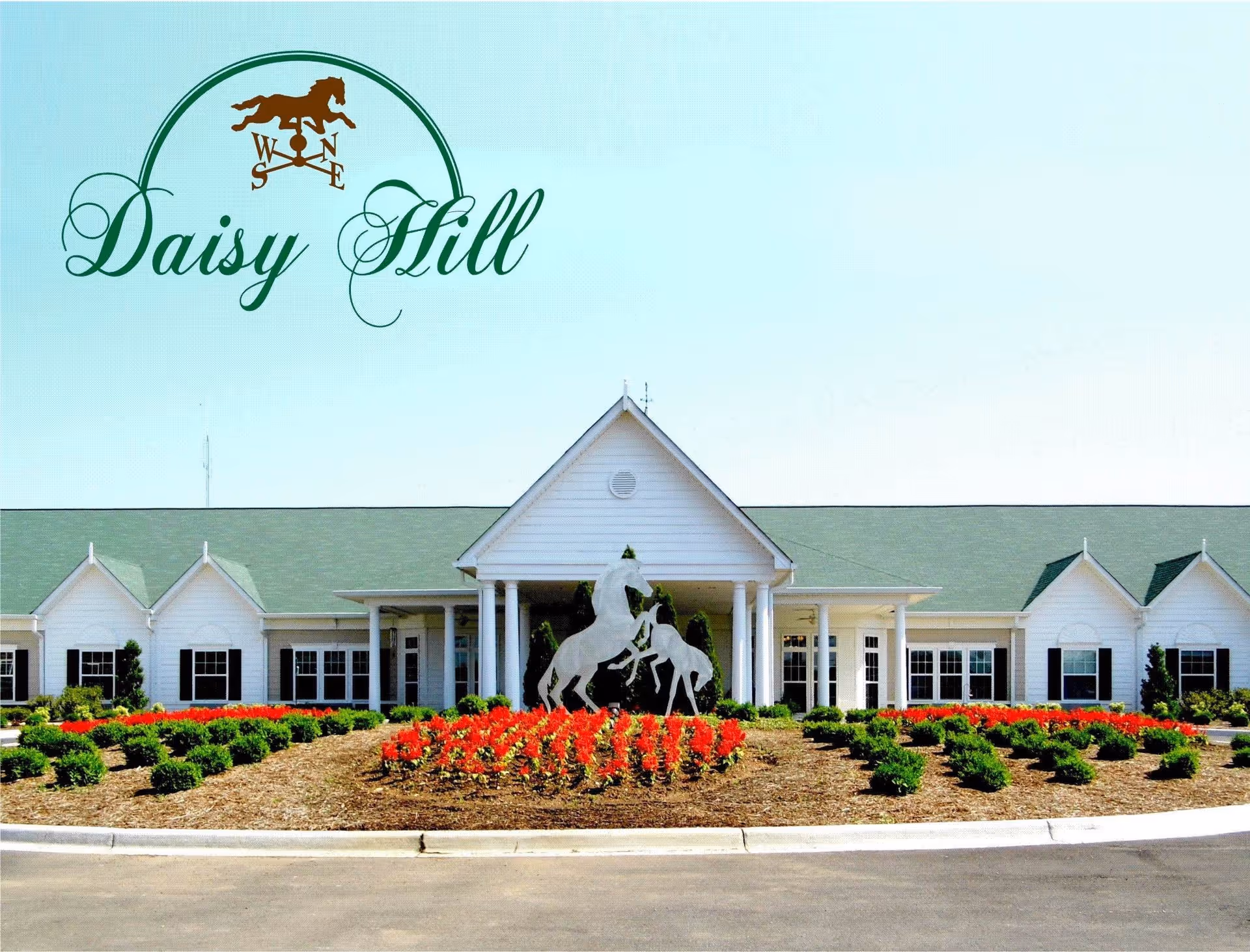 Front exterior view of Daisy Hill Senior Living facility featuring a white building with a green roof, a central entrance with columns, a landscaped garden with red flowers and green shrubs, and a sculpture of two horses in the center.