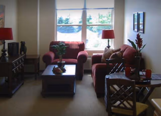 A cozy living room with two red upholstered armchairs and a matching sofa arranged around a wooden coffee table. There are two red lamps on side tables, a small dining table with two chairs, and a large window letting in natural light. The room is decorated with plants and framed pictures on the wall.