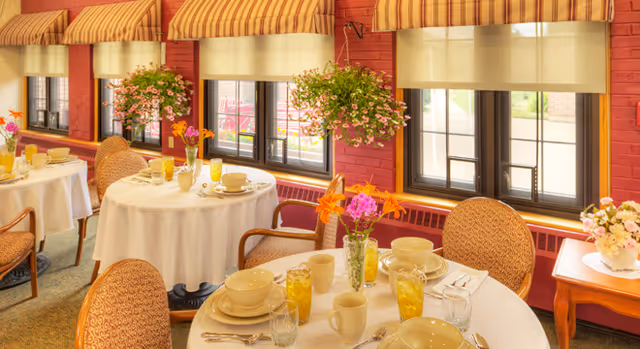 A bright dining room with round tables covered in white tablecloths, set with beige dishes, glasses of iced tea, and silverware. The room features large windows with striped valances and hanging flower baskets. Comfortable upholstered chairs surround the tables, and a small wooden side table with a floral arrangement is visible near the windows.