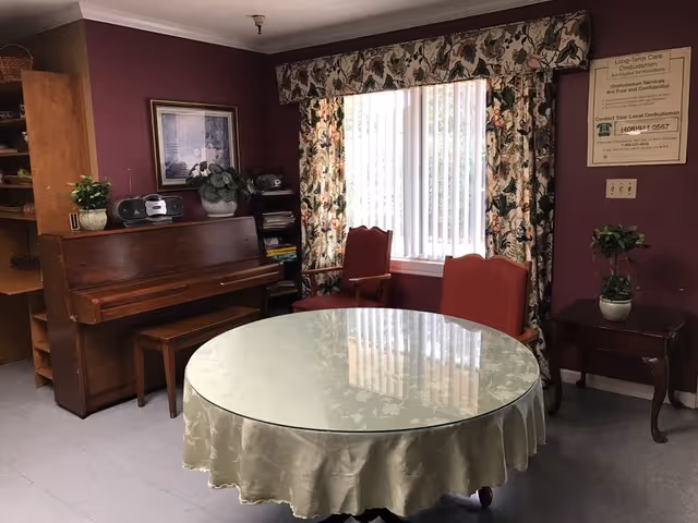 Common room with a round glass-covered table, red chairs, an upright piano, and a window with floral curtains.