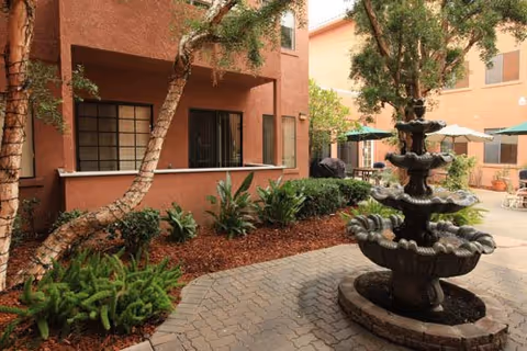 Outdoor courtyard area with a multi-tiered stone fountain in the foreground, surrounded by paved walkways, green plants, and trees. The courtyard is enclosed by a two-story building with windows and patio umbrellas providing shaded seating areas.