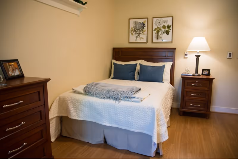 A cozy bedroom with a wooden bed frame, white quilted bedspread, two blue pillows, and a folded gray blanket on the bed. There is a wooden nightstand with a lamp, a small clock, and a framed photo. A wooden dresser with a framed photo is on the left side. Two botanical prints hang on the wall above the bed.