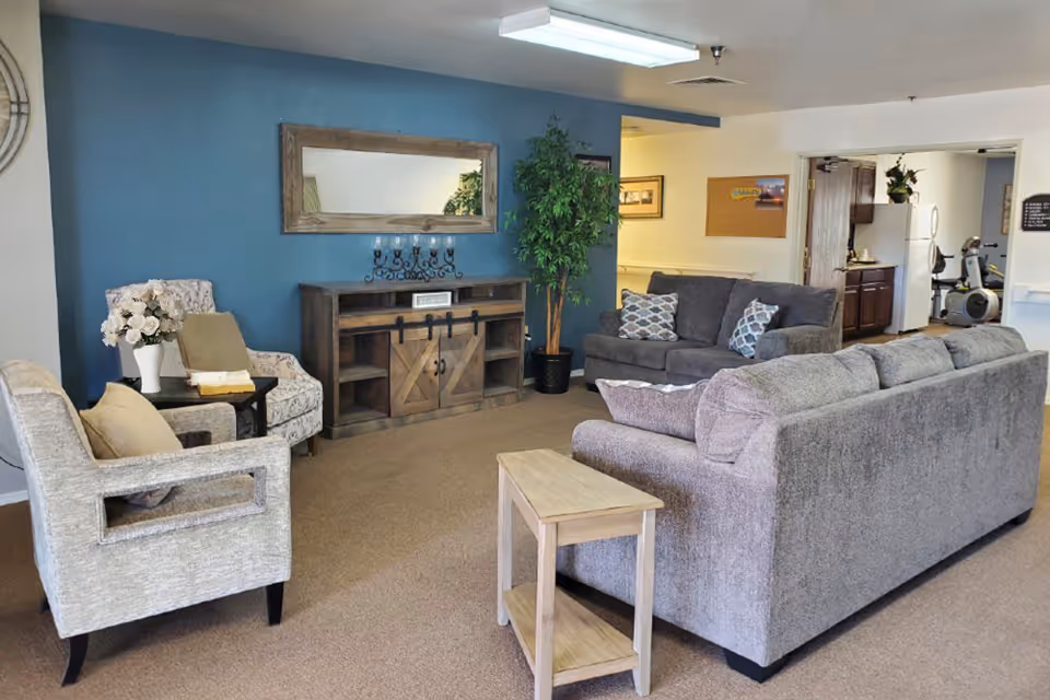 A cozy living room area with two gray sofas, two patterned armchairs, a small wooden side table, a wooden cabinet with barn-style sliding doors, a large rectangular mirror on a blue accent wall, a potted plant, and a vase of white flowers on a side table. In the background, there is a kitchen area with a refrigerator and exercise equipment visible.