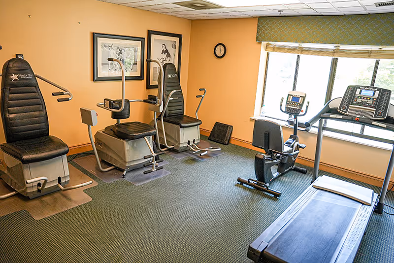 A small fitness room with exercise equipment including two seated strength training machines, a recumbent stationary bike, and a treadmill. The room has beige walls, a green carpet, two framed pictures on the wall, a clock, and a window with a view outside.