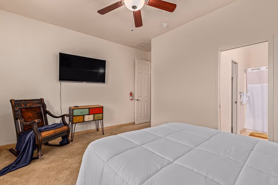 Bedroom with a bed in the foreground, wall-mounted TV, wooden rocking chair, small colorful dresser, ceiling fan, and an open door leading to a bathroom.