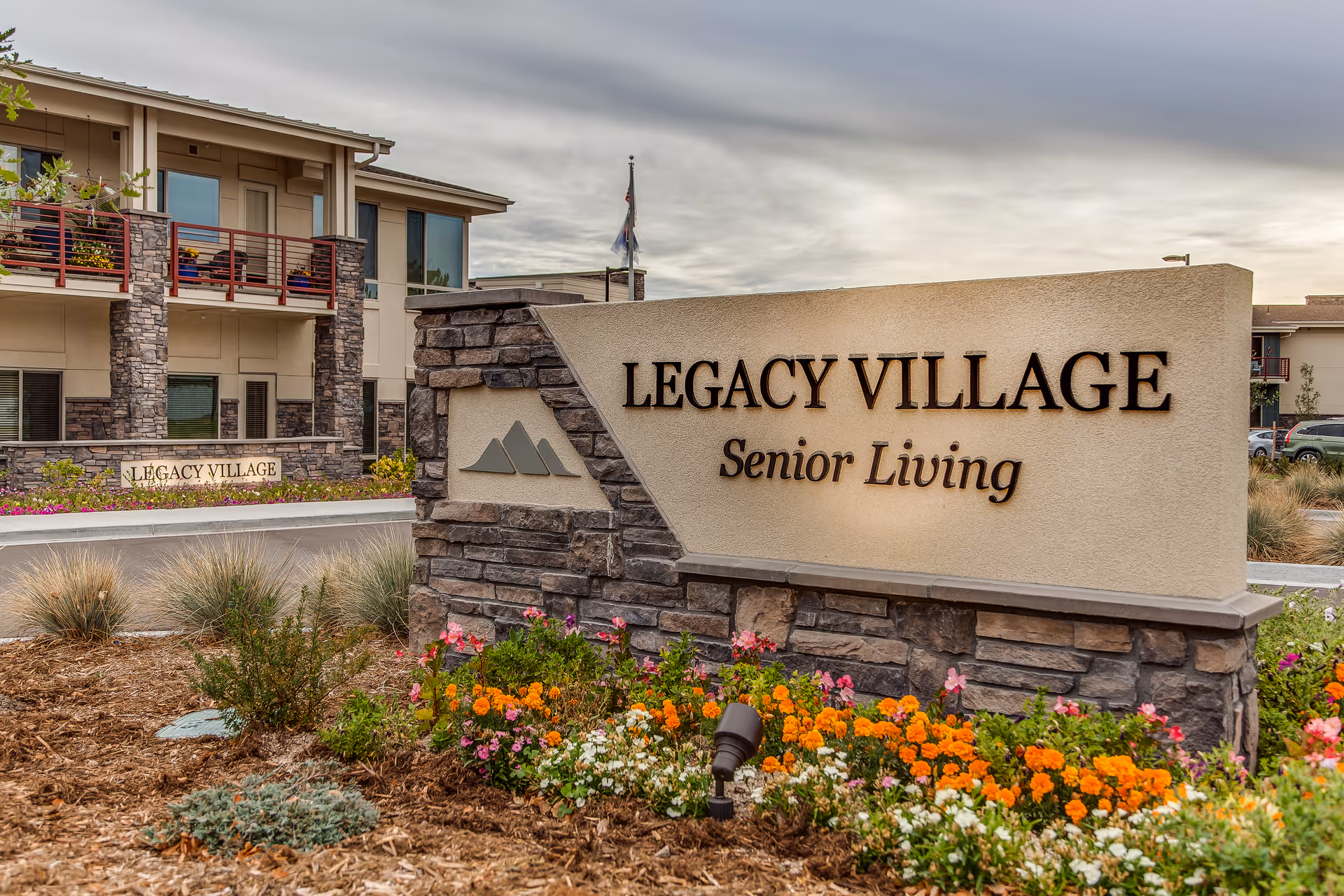 Stone and stucco entrance sign reading "Legacy Village Senior Living" with flowers and the residence building behind it.