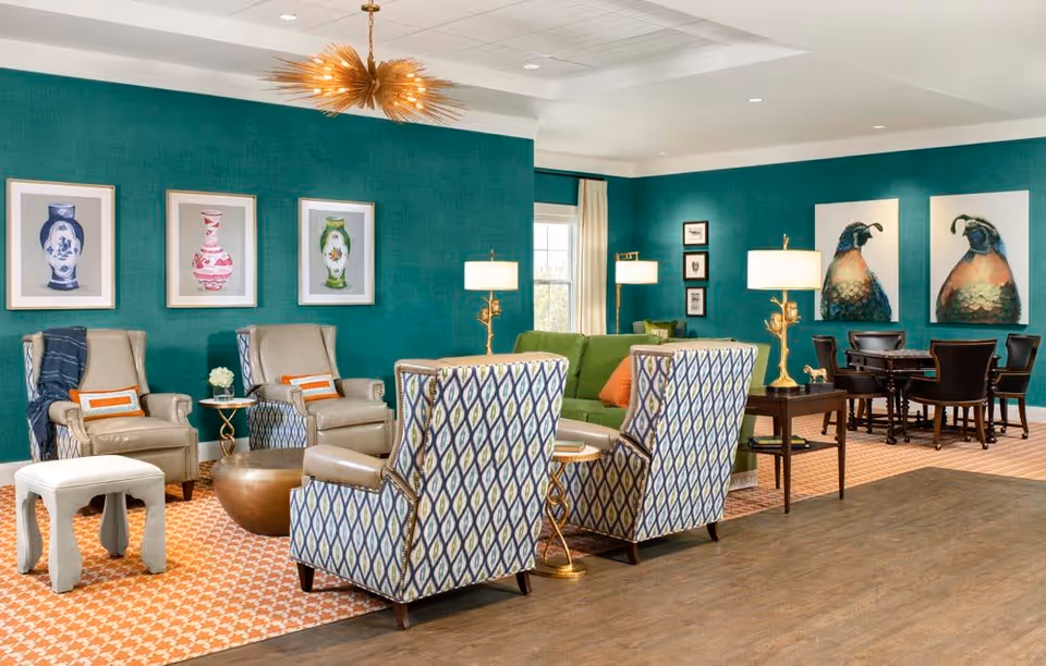 A bright and spacious living room area with teal walls adorned with framed artwork of vases and birds. The room features a green sofa, patterned armchairs, beige armchairs with orange accent pillows, a small white stool, and several side tables with lamps. In the background, there is a dark wood table with chairs arranged around it.