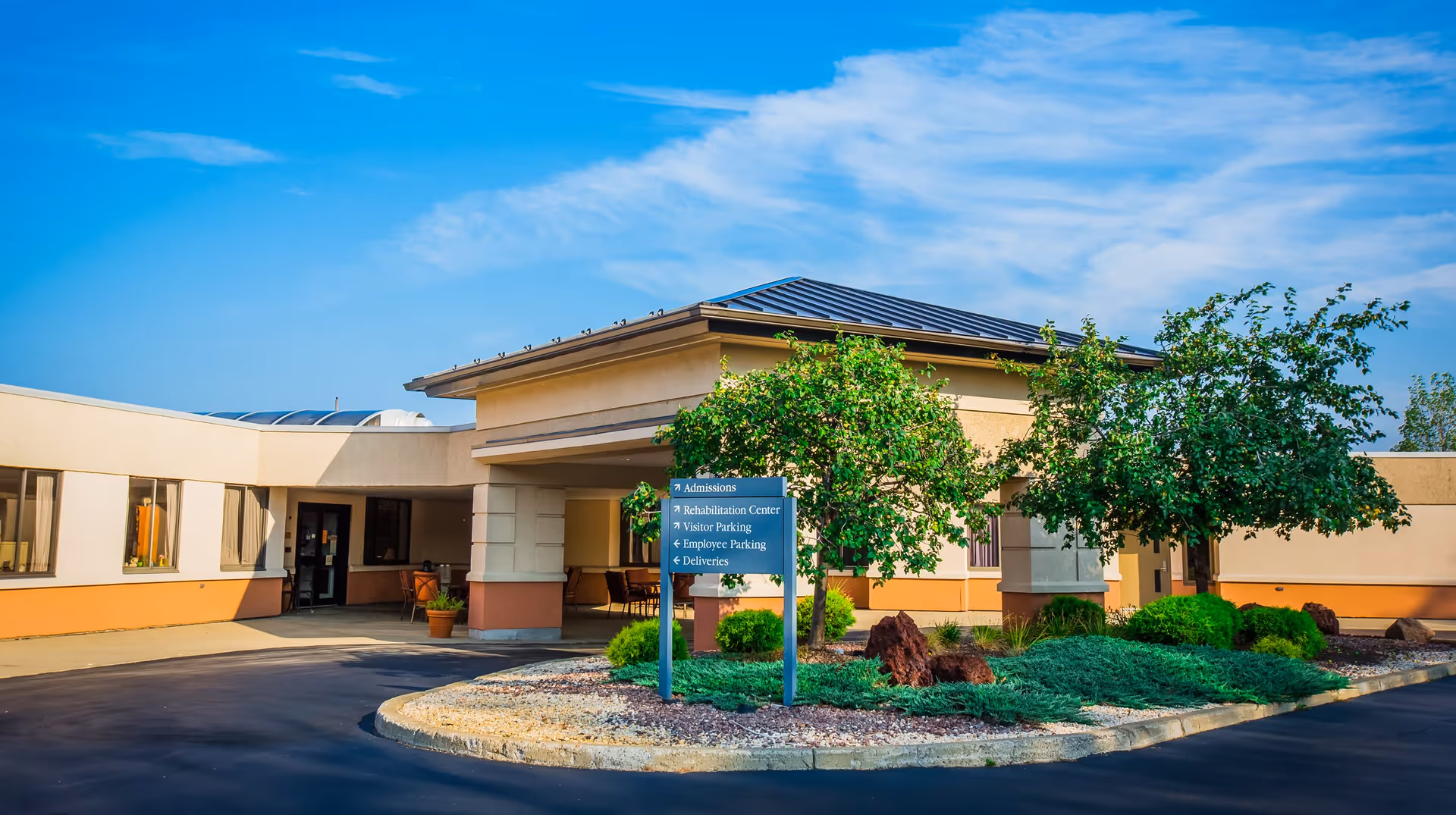 Front entrance of a single-story health care building with a covered porte-cochere, driveway, sign, and landscaped trees and shrubs.