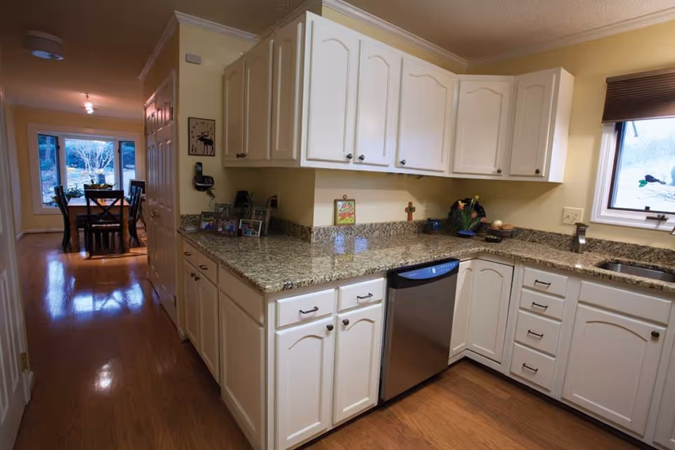 A kitchen with white cabinets, granite countertops, a stainless steel dishwasher, and a sink under a window. The kitchen opens into a hallway leading to a dining area with a table and chairs near large windows.