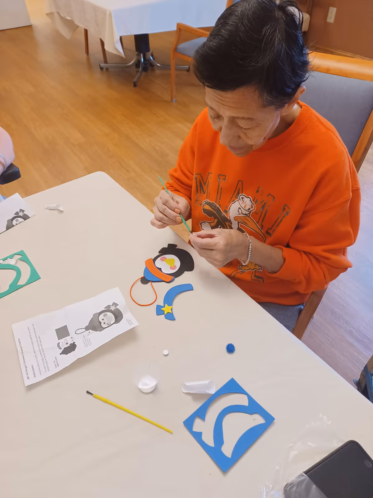 An elderly person wearing an orange Miami sweatshirt is sitting at a table engaged in a craft activity, assembling colorful paper pieces and using a small paintbrush. Various craft materials and instructions are spread out on the table.