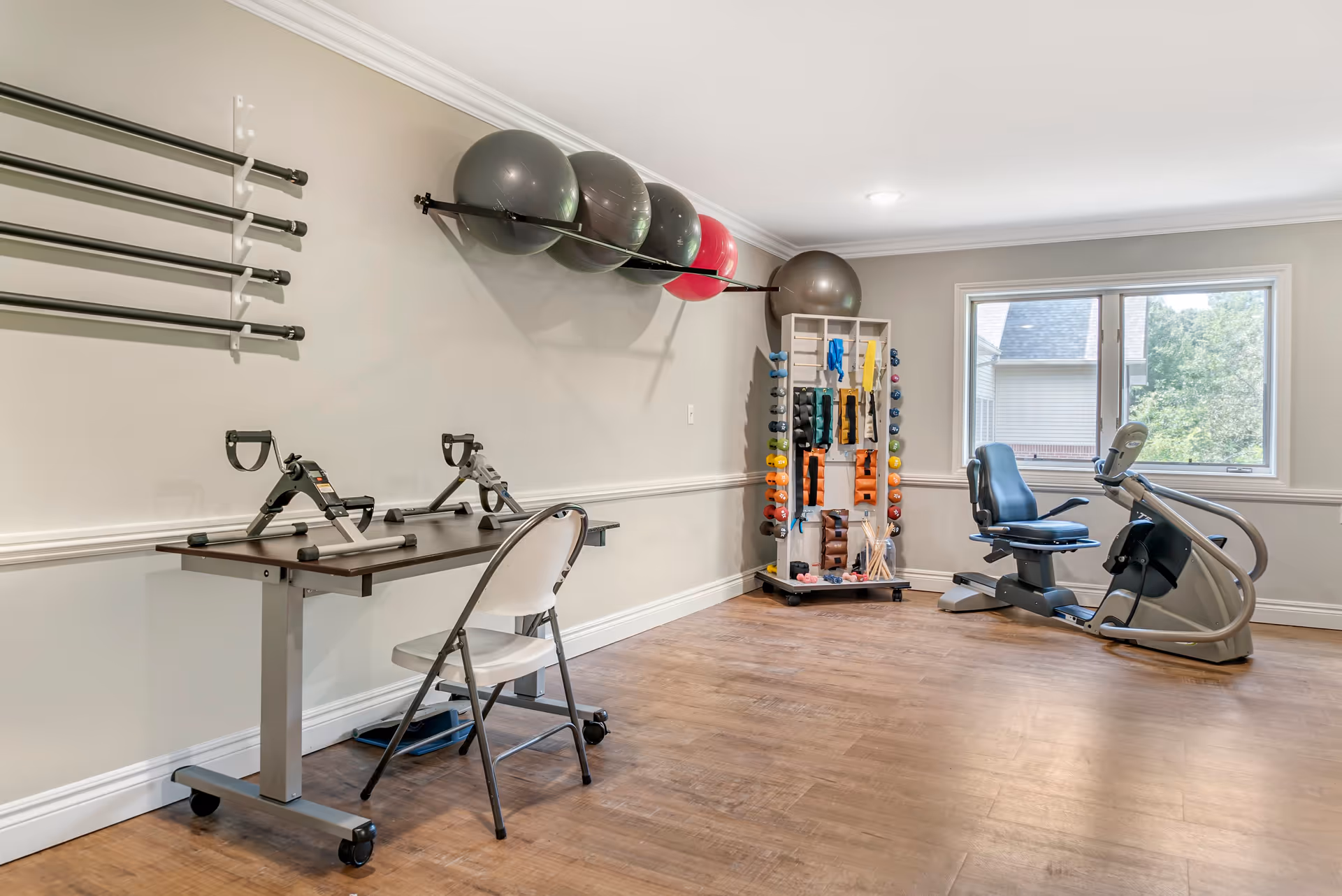 A fitness room with exercise equipment including a recumbent bike, a rack of colorful dumbbells, resistance bands, exercise balls mounted on the wall, and two hand pedal exercisers on a table with a folding chair. The room has wooden flooring, light-colored walls, and a window letting in natural light.