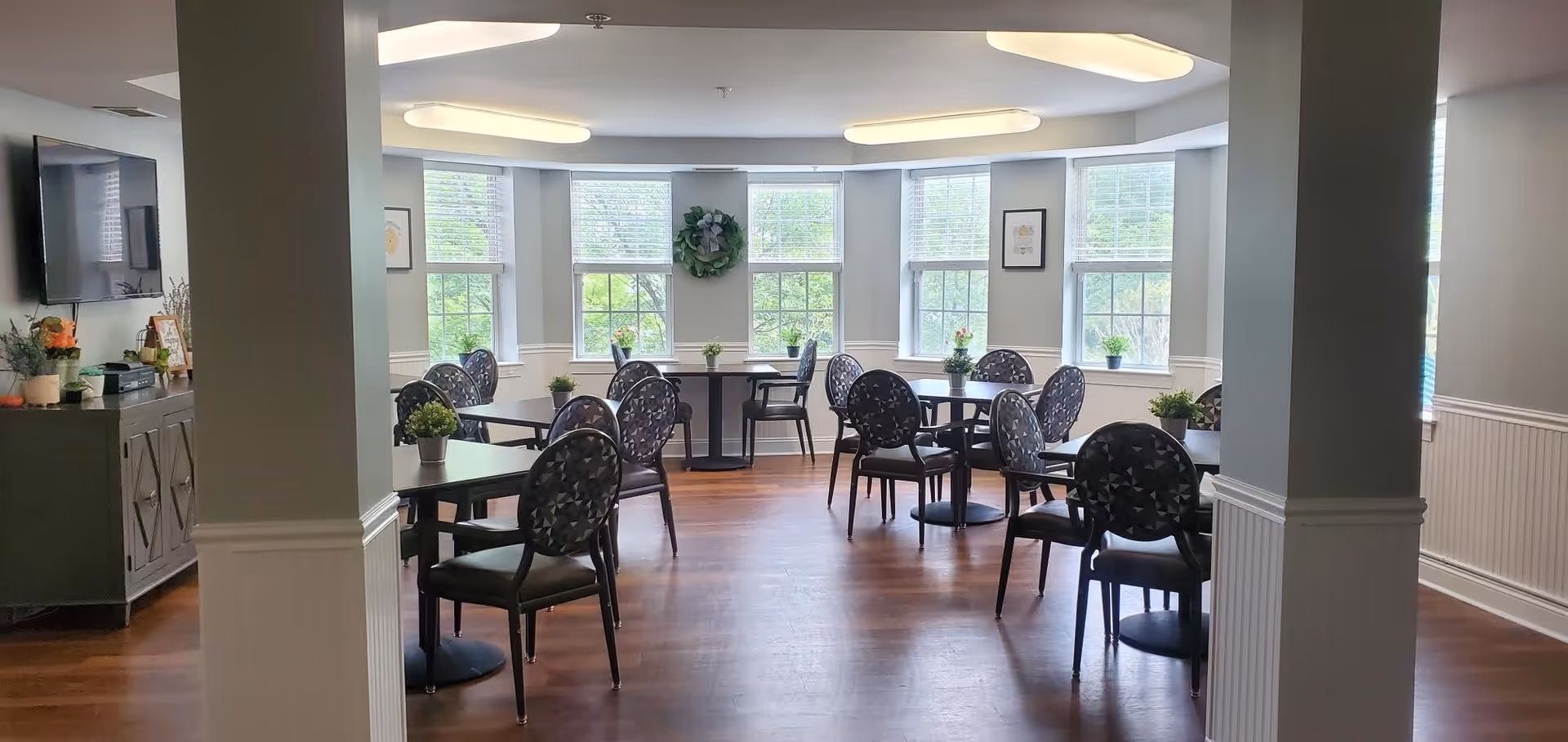 A bright dining room with multiple round tables and black chairs arranged neatly. The room has large windows letting in natural light, light gray walls, and wooden flooring. There are small potted plants on each table and a wreath hanging on the wall between the windows. A TV is mounted on the left wall above a cabinet with decorative items.