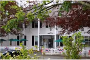 Front entrance of a two-story white nursing home with a covered porch, picket fence decorated with bunting, and green patio umbrellas framed by trees.
