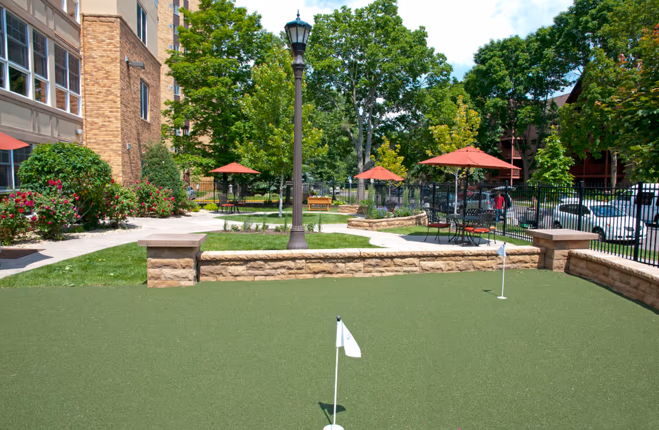 Outdoor area at The Alton featuring a small putting green with two white flags, surrounded by stone walls. There are several red patio umbrellas with tables and chairs on paved walkways. The area is landscaped with green grass, bushes, and trees, and a tall lamppost stands near the center. A brick building is visible on the left side, and a black metal fence encloses the space with parked cars beyond it.
