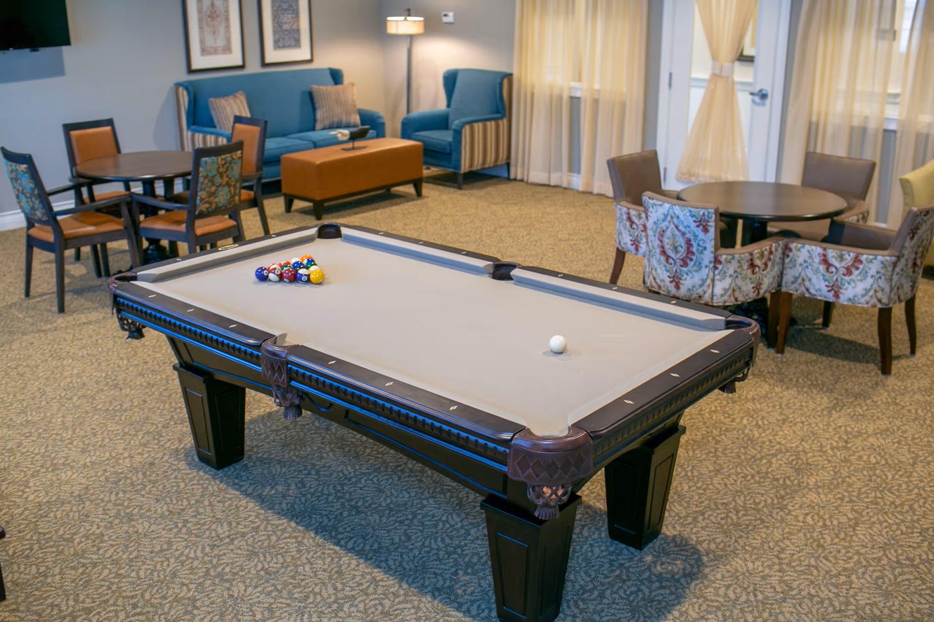 A senior living facility common area featuring a pool table with billiard balls arranged for a game. Surrounding the pool table are several seating areas with round tables and chairs, a blue couch, an armchair, and a brown ottoman. The room is carpeted and has large windows with sheer curtains allowing natural light to enter.