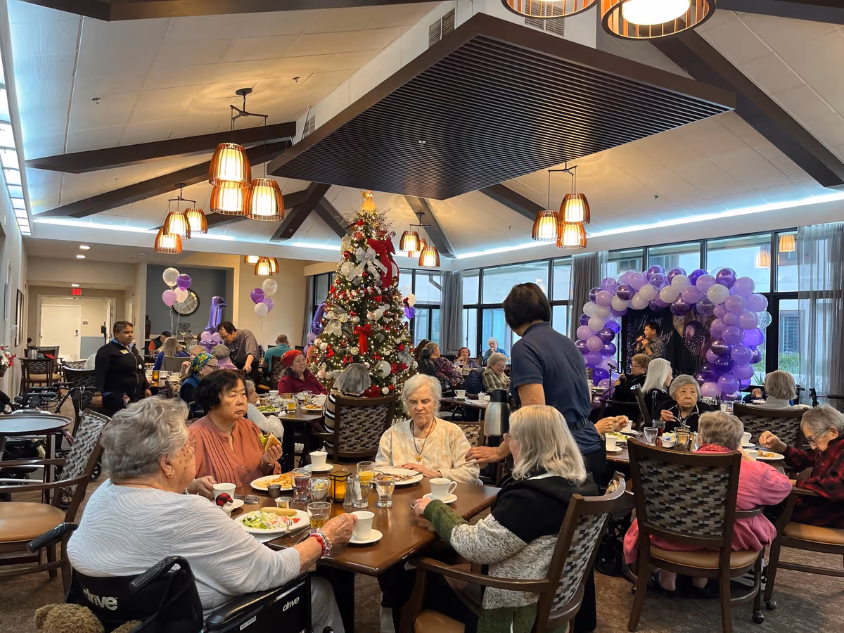 A festive dining room in a senior living facility decorated for a celebration with a Christmas tree and purple and white balloon arch. Several elderly residents are seated at tables eating and socializing, while staff members assist them. The room has large windows, warm lighting, and a high ceiling with exposed beams.